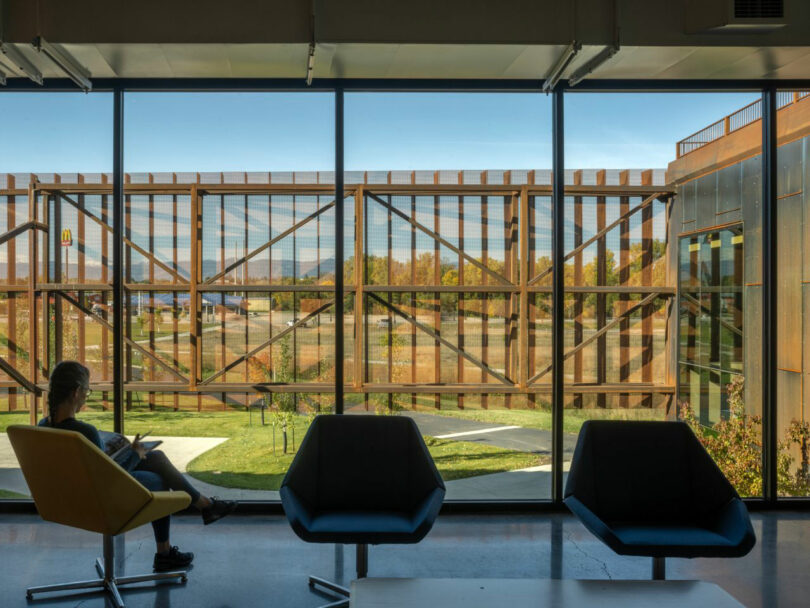 A person sits on a yellow chair reading a book in a modern building designed by CLB Architects. Large windows reveal exterior rusty metal beams and a landscape with trees and a clear sky.