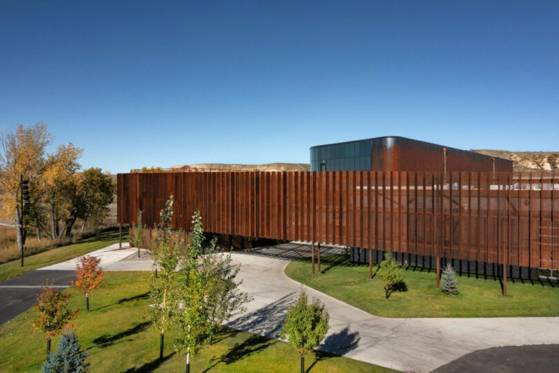 Designed by CLB Architects, the modern building features a rust-colored facade, harmoniously set amidst lush trees and grass under a clear blue sky.