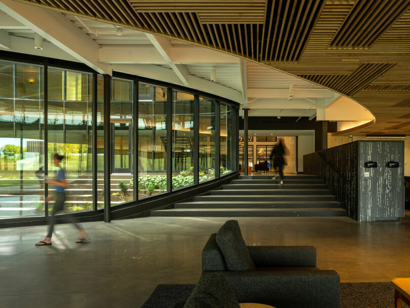Designed by CLB Architects, the modern office interior boasts large windows with lush plants visible outside. In the foreground, a sleek gray couch complements the scene as two blurred figures walk through this harmonious space.