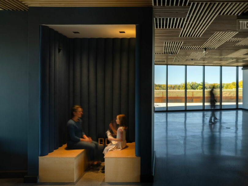 Two people sit across from each other in a dark alcove seating area, designed with CLB Architects' signature modern touch. Another person walks by large windows on the right, highlighting the room's elegant wood accents.