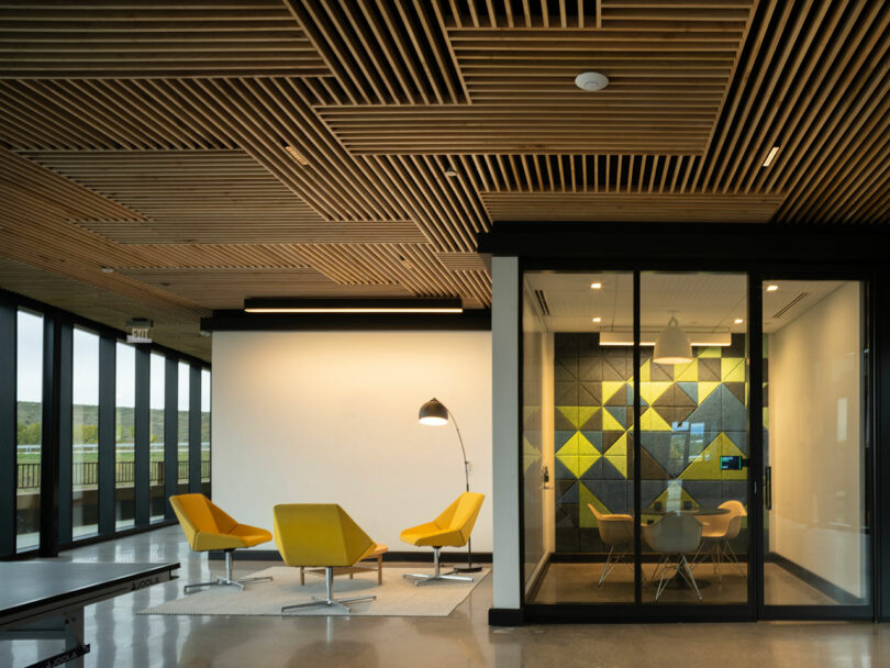 Modern office space by CLB Architects features yellow chairs, a floor lamp, and patterned acoustic panels. Large windows and a wooden ceiling elevate the natural light and ambiance.