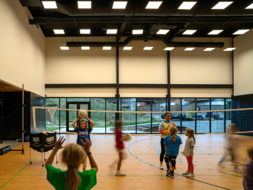 Kids enthusiastically play volleyball indoors, coached under CLB Architects' beautifully designed space, featuring a polished wooden floor, expansive windows, and bright overhead lights.