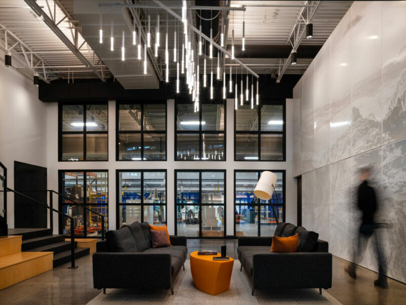 The modern office lobby, designed by CLB Architects, features two dark sofas and an orange coffee table under sleek pendant lights. A blurred person walks by as workshops are visible through the windows, illuminated softly by a white floor lamp.