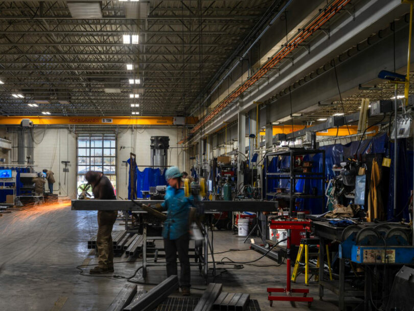 In an industrial workshop scene designed by CLB Architects, workers expertly operate equipment amidst the welding of steel beams. The space is illuminated by overhead lighting, while shelves neatly line the walls, showcasing a blend of functionality and architectural finesse.