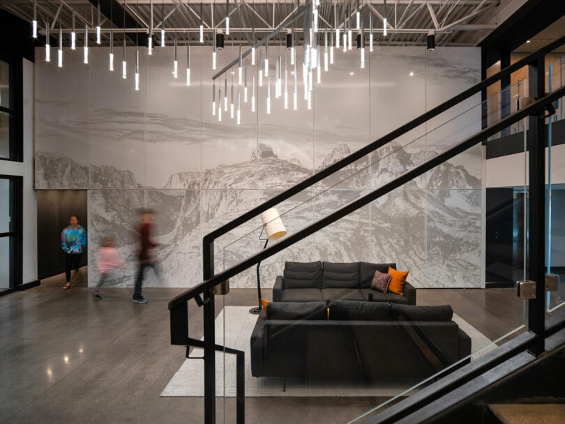 Designed by CLB Architects, this modern interior features a large mountain mural, a sleek black sofa, and a contemporary chandelier. The scene comes alive with two people in motion, while a striking metal staircase sets the stage in the foreground.