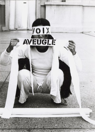 A woman dressed all in white is crouched down on a sidewalk. Her eyes and mouth are covered with a white piece of cloth that reads 'VOIX AVEUGLE'.