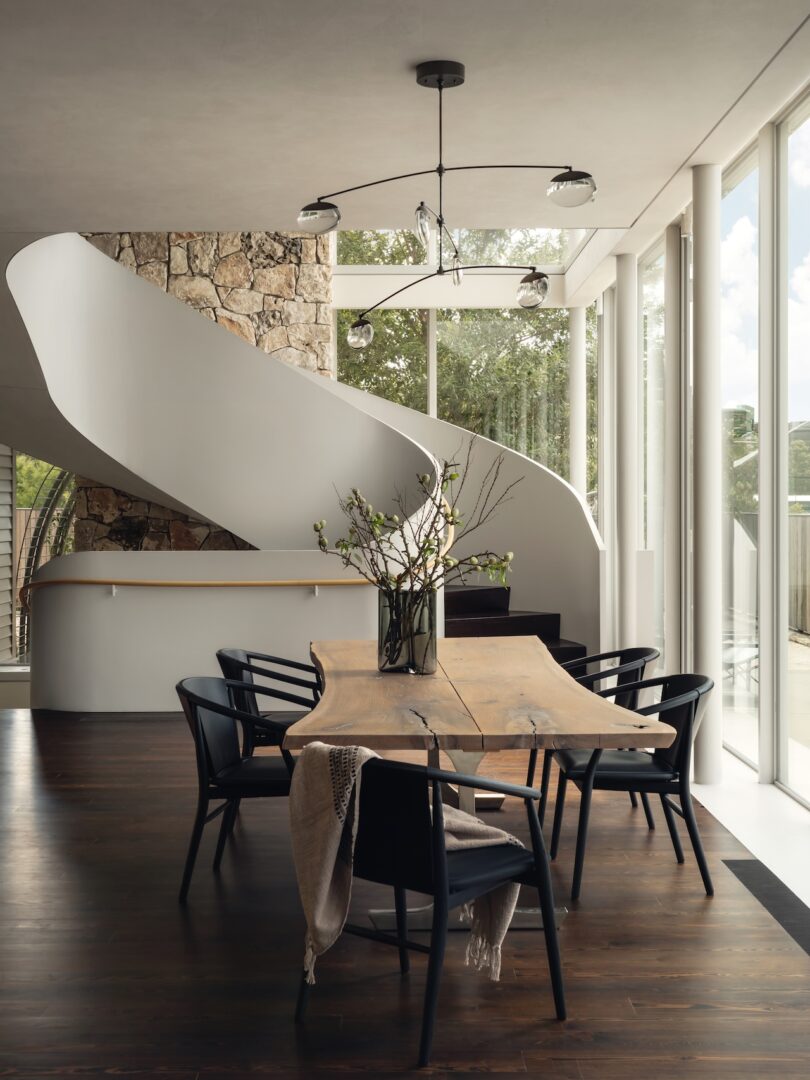 Modern dining room with a wooden table, black chairs, and a spiral staircase. Large windows let in natural light, highlighting the stone wall and minimalist decor.
