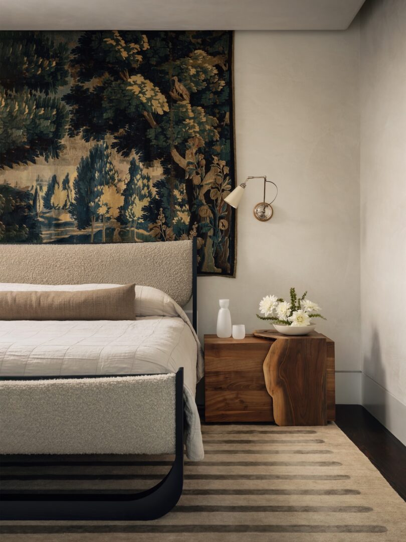 Bedroom with a bed, wooden nightstand, and rug. A tapestry with trees hangs above the bed. A table lamp, white carafe, and flowers are on the nightstand. Walls are light-colored.