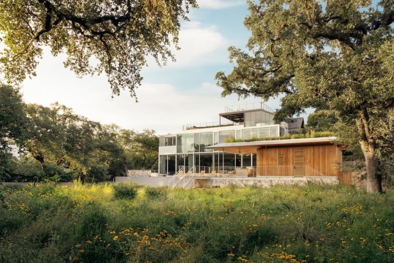 Modern glass and wood house surrounded by trees and a grassy field under a blue sky.