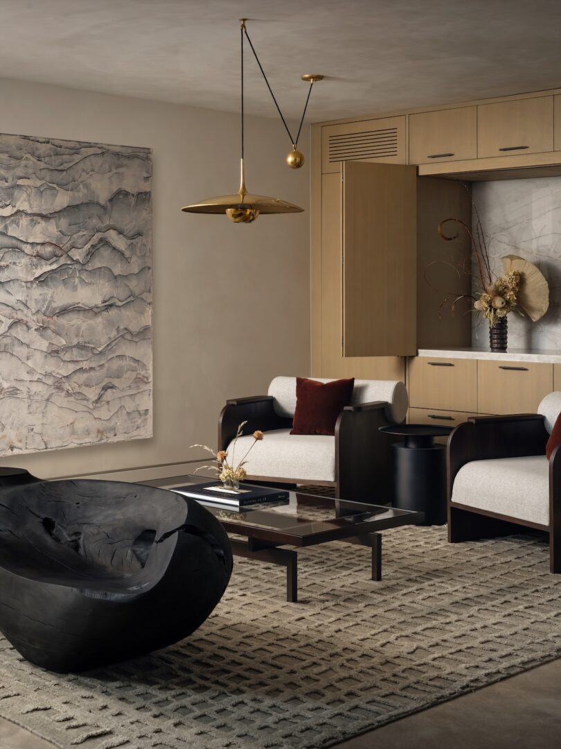 Modern living room with abstract wall art, a black coffee table, two beige armchairs with brown cushions, a sculptural chair, and a brass pendant light over a textured rug.