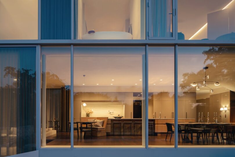 Exterior view of a modern building with large glass windows, revealing a lit-up interior with dining and living areas. Evening setting with reflections of trees in the glass.
