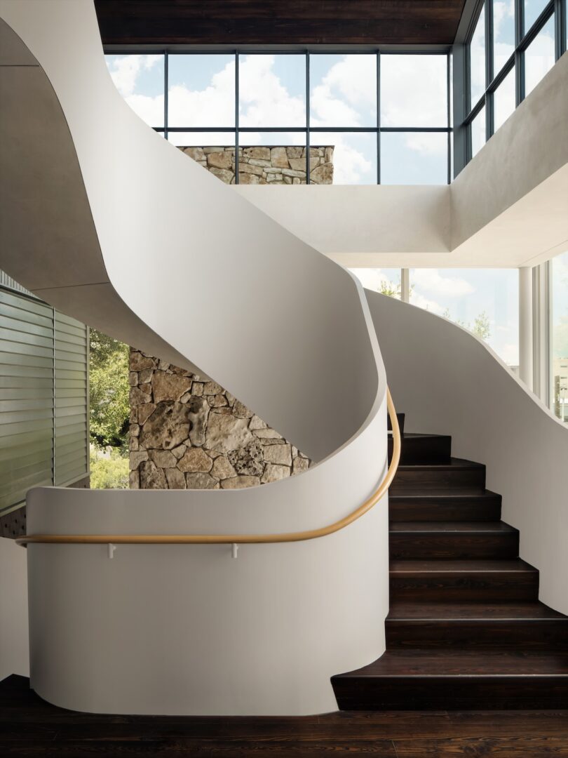 A modern, curved staircase with white walls and a wooden handrail leads up to the second floor. Large windows provide natural light, and a stone wall adds texture.