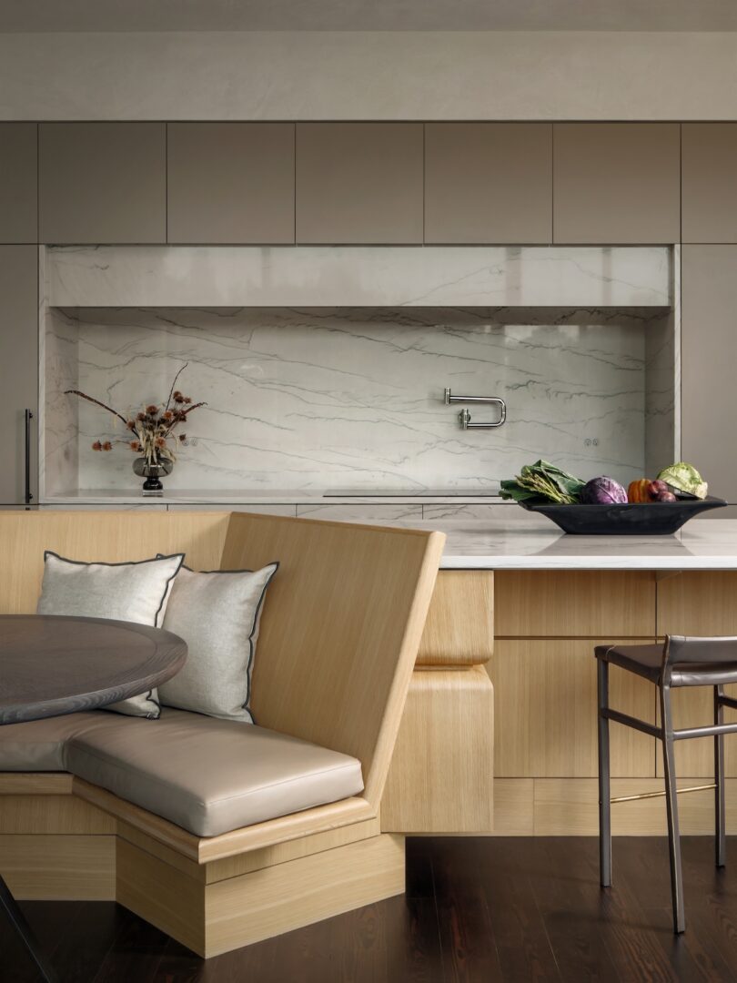Modern kitchen with beige cabinets, marble backsplash, built-in bench seating, round table, and bar stool. A black bowl with assorted vegetables is placed on the counter.