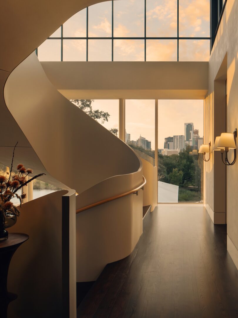 A modern interior with a curved staircase, large windows, and a view of a city skyline against a sunset. The walls are light-colored with a sconce, and there's a table with decorative items.