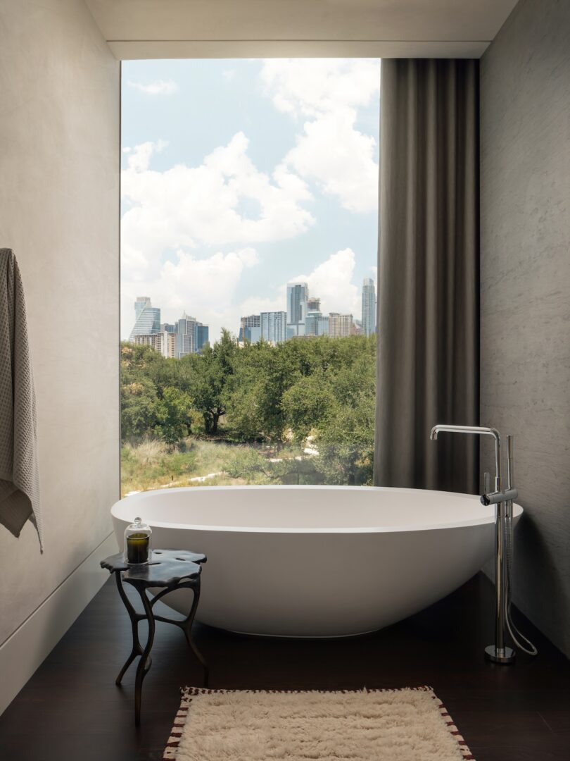 Freestanding bathtub near a large window with a city skyline view. A small table with a candle and a hanging towel are nearby.