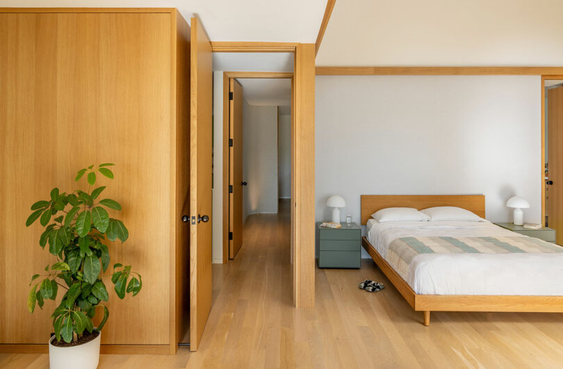Minimalist bedroom with a wooden floor, a large bed, green bedside tables, a potted plant, and open wooden doors leading to a hallway.