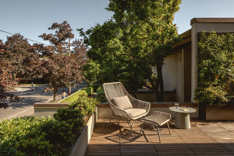 A cozy outdoor deck with a wicker chair, ottoman, and small table holding a glass. Surrounded by lush trees and a view of a cyclist on the street below.