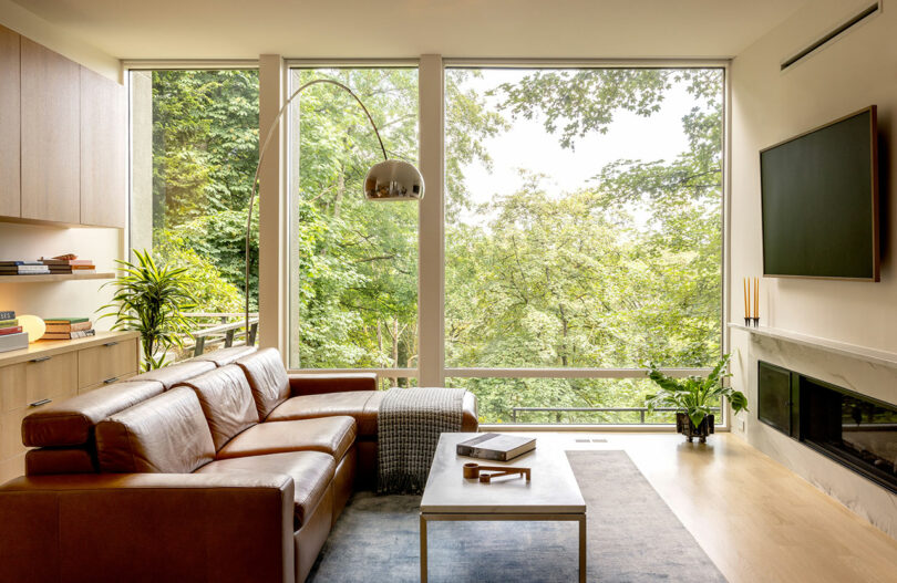 A modern living room with a brown leather sofa, large windows showcasing a tree view, a floor lamp, a TV mounted above a fireplace, and a wooden coffee table with books.