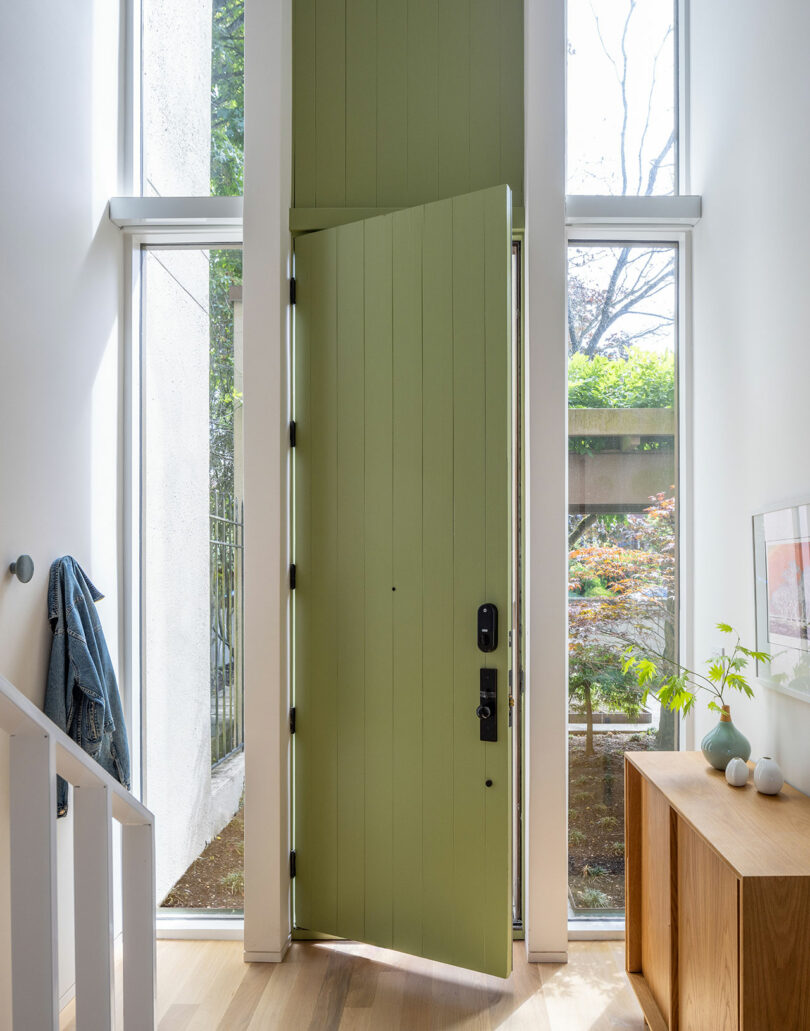 A tall, light green door is partially open, set between large windows in a modern interior entryway with light wood flooring and a small wooden table with decorative items.