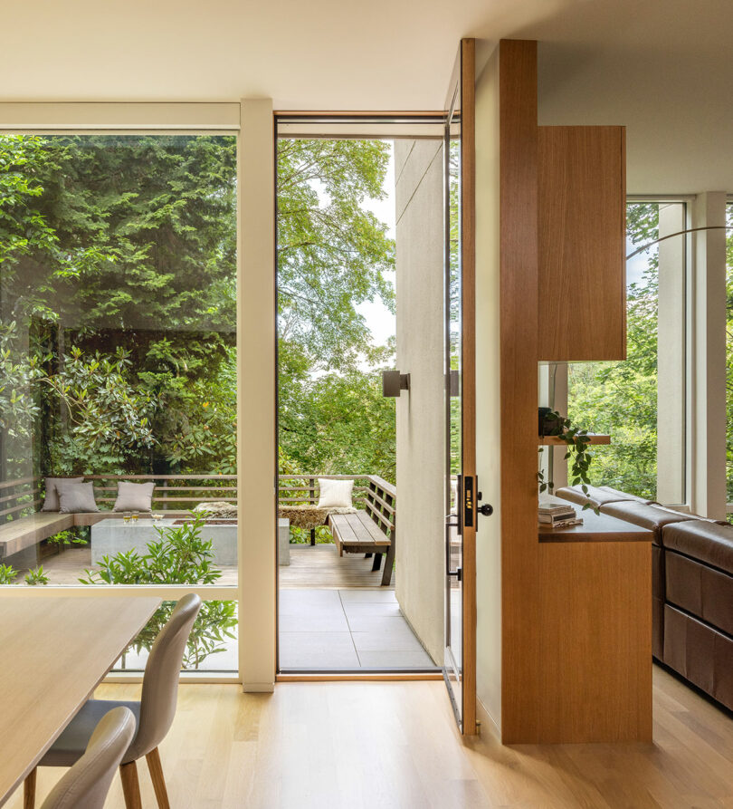 Modern interior with large glass doors leading to a patio. Light wooden floors, dining table, chairs, and brown sofa are visible. Greenery and outdoor seating are seen through the open door.