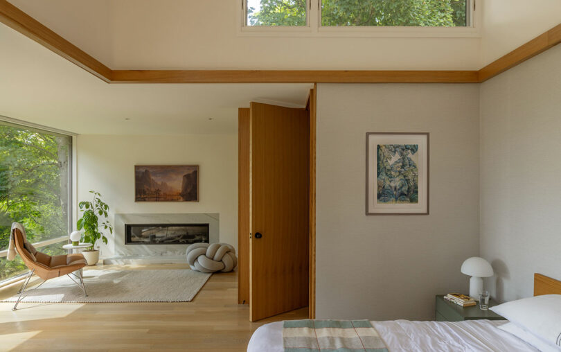 Modern bedroom with a bed, a chair near a large window, a knot cushion on the floor, a fireplace, and framed artwork on the walls. Neutral tones and natural light create a serene atmosphere.