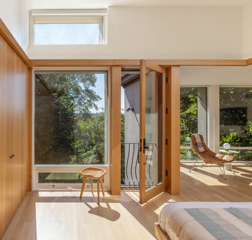 Modern bedroom with large windows, a wooden chair, and a leather lounge chair. Sunlight streams in, highlighting the wooden floor and white walls. Bed partially visible in the foreground.