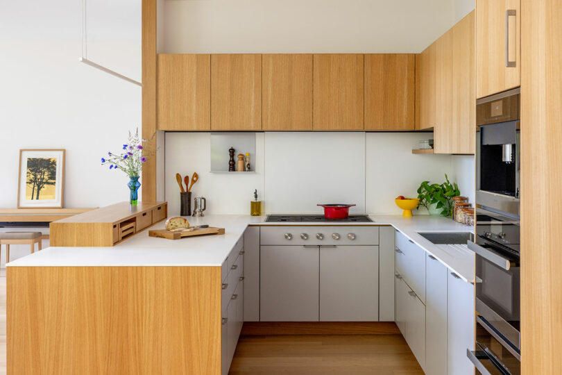 Modern kitchen with light wood cabinets, white counters, and stainless steel appliances. A red pot on the stove, bread on a cutting board, and colorful flowers in a vase.