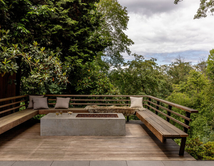 Outdoor wooden deck with L-shaped bench, cushions, and a concrete fire pit in a lush, forested setting under cloudy skies.