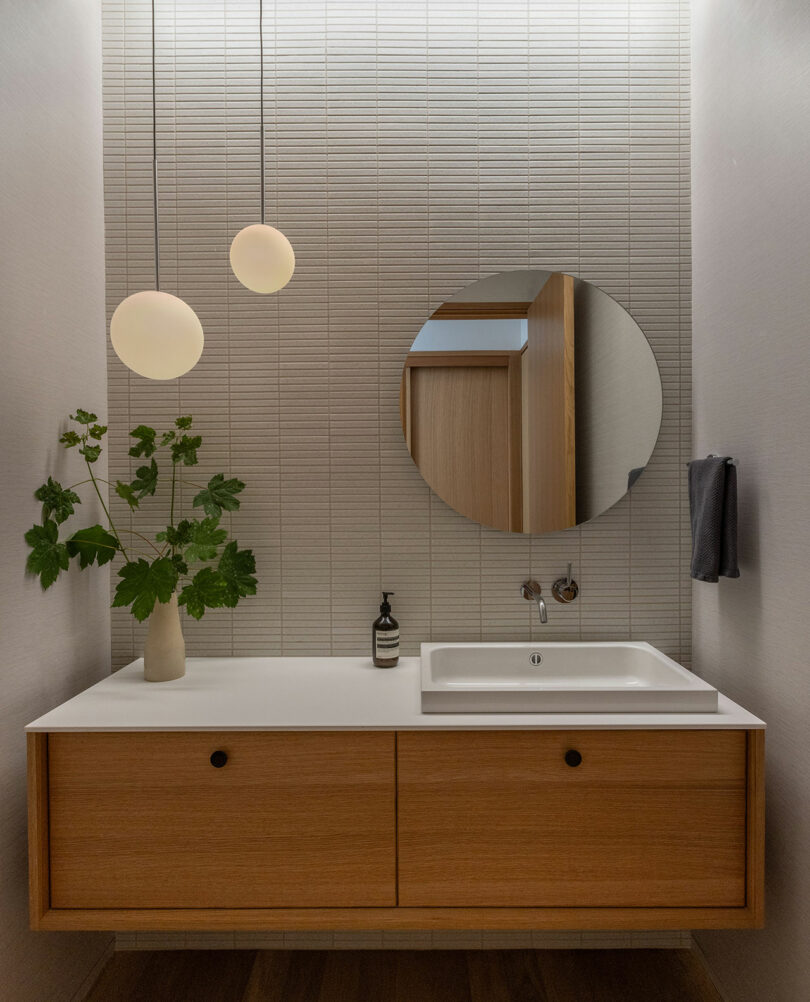 Minimalist bathroom features a wooden vanity with a rectangular sink, round mirror, hanging globe lights, and a potted plant, against a textured tiled wall.