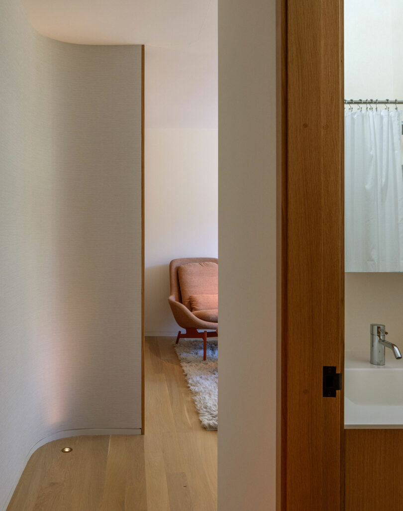 A narrow hallway leads to a room with a brown armchair and a textured white rug. A section of a bathroom with a faucet and shower curtain is visible on the right.