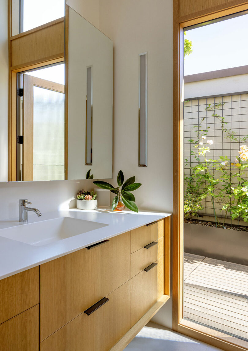 A modern bathroom features a wooden vanity with a white countertop and a rectangular mirror. A plant sits near the sink. Sunlight streams in from a large window overlooking a garden.