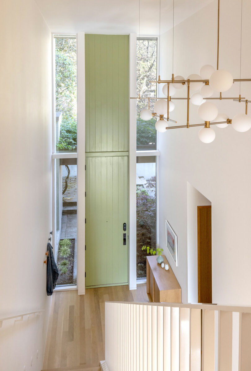 A modern, minimalist entryway with a tall green door, white walls, wooden flooring, a brass chandelier, and a console table with decor. A coat hangs on a hook by the door.