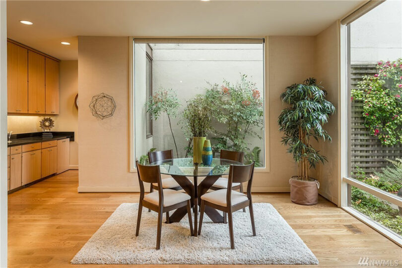 Minimalist dining area with a round glass table and four chairs on a rug. Large window reveals greenery outside. Light wooden floor and cabinets in the open kitchen space.
