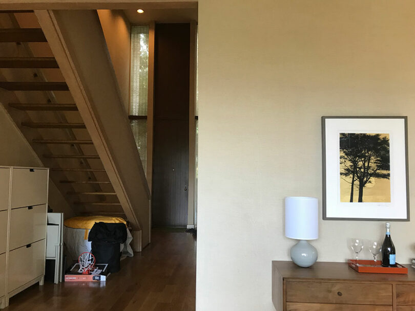 A minimalist room with wooden floors features a staircase, a bed, a dresser with a lamp, wine bottle, and glasses, and a wall picture of a tree.
