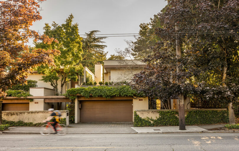 A cyclist rides past a modern house with a two-car garage, surrounded by trees and greenery, under a clear sky.