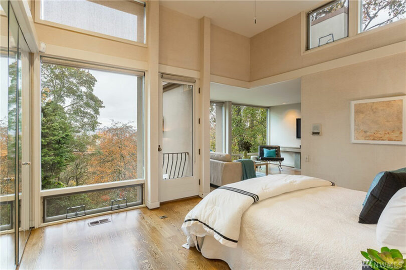 Bedroom with large windows, a bed with white linens, and a small seating area. Walls are light-colored, and there's a view of trees outside. Artwork hangs above a writing desk.