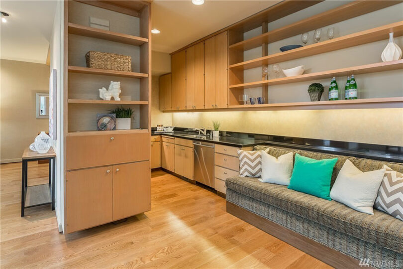 Modern kitchen with wooden cabinets, stainless steel appliances, open shelving, and a built-in cushioned bench with patterned pillows on a wooden floor.