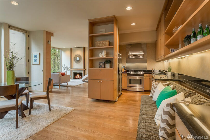 Modern open-concept living area with hardwood floors, a dining table, a fireplace, and a kitchen featuring stainless steel appliances. Sofa and shelving provide additional storage and seating.