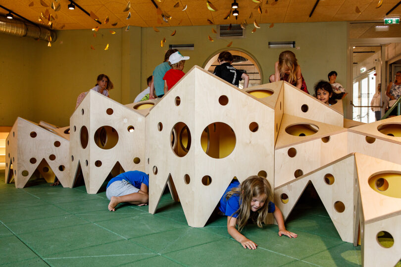 Children crawl and play through a wooden, cheese-shaped tunnel in an indoor play area with a green floor.