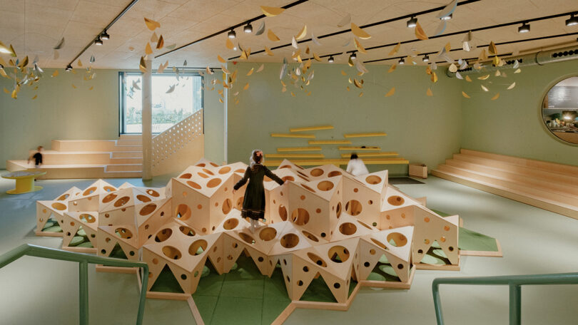 Children play on a geometric climbing structure in a spacious, modern indoor play area with green and yellow decor.