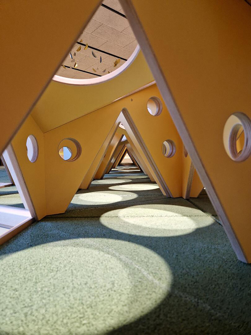 Triangular wooden structures with circular cutouts are aligned in a row, casting shadows on a green carpeted floor.