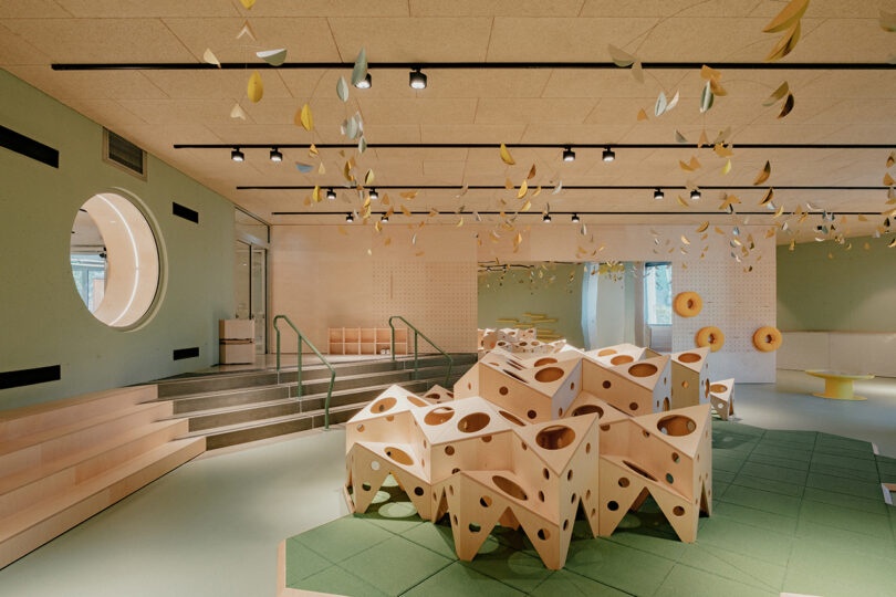 Modern children's playroom with geometric wooden climbing structures, suspended leaf decorations, and a circular window. The room has a green and beige color scheme with soft lighting.