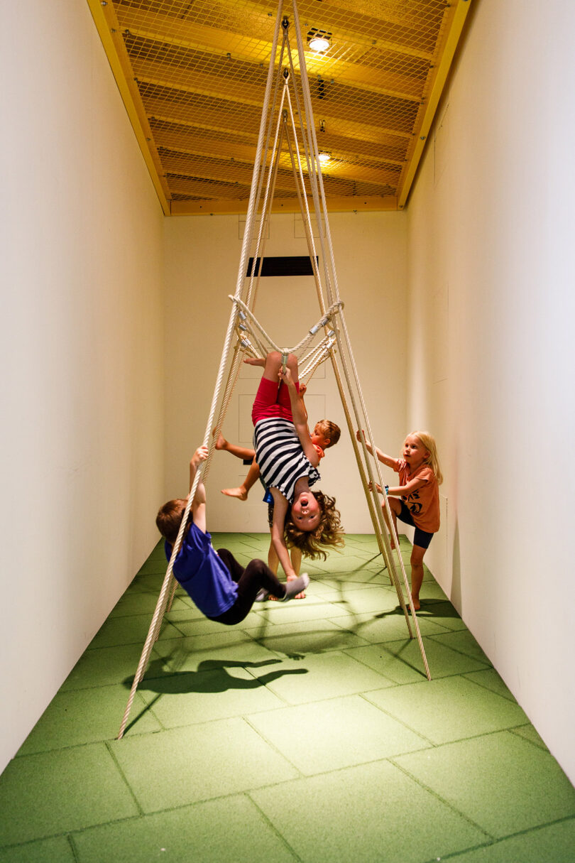 Children playing and hanging on a rope pyramid structure in a narrow room with green flooring and a yellow ceiling.