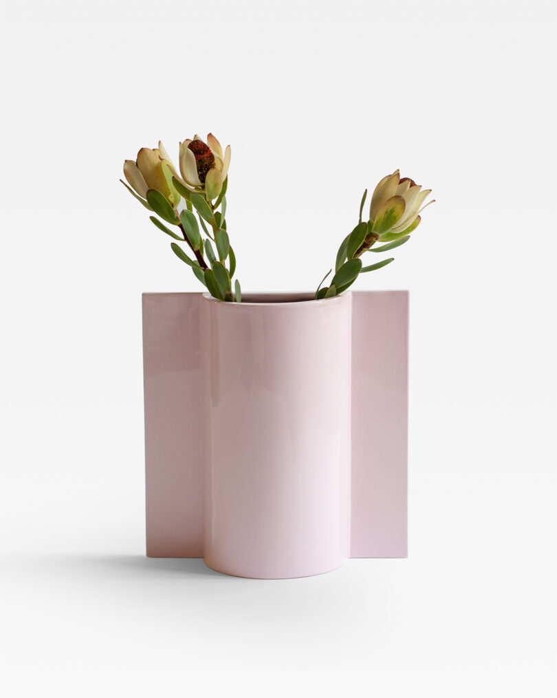 A glossy pink vase with geometric shapes holding two light-colored flowers with green leaves against a plain white background.