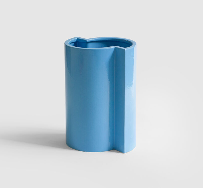 A modern, glossy blue ceramic vase with a cylindrical shape and a geometric protrusion on one side, set against a plain light background.