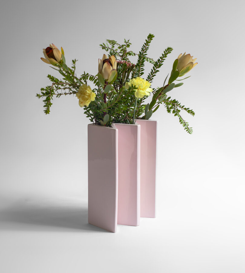A modern, pink ceramic vase with three vertical sections holds a bouquet of yellow flowers, greenery, and large closed buds against a plain background.