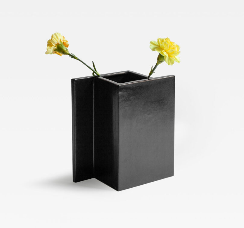 A Kish O Mat Sara Khamisi-inspired black rectangular vase with two vibrant yellow flowers elegantly extending from the top, set against a plain white background.