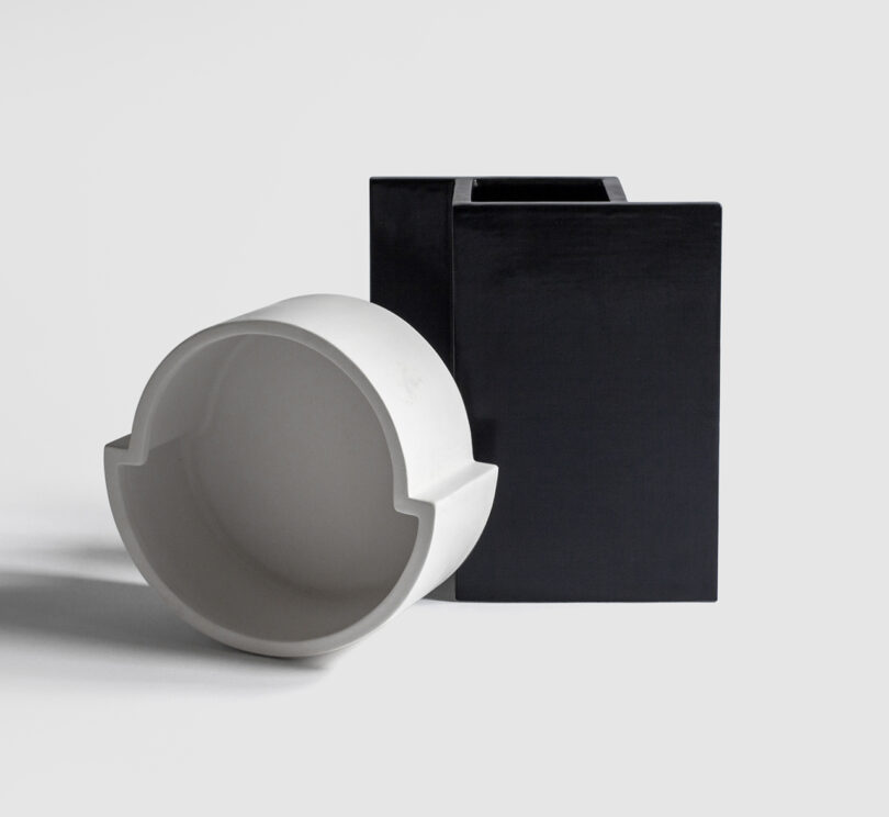 A white, round object with cutouts is positioned partially inside a black, square hollow block against a plain background.