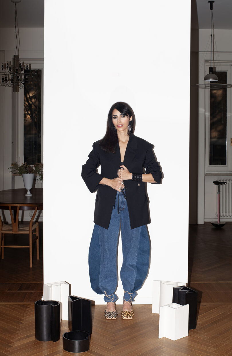 Person in a black blazer and loose jeans stands against a white backdrop, surrounded by geometric-shaped objects, in a room with wooden flooring and a dining area.