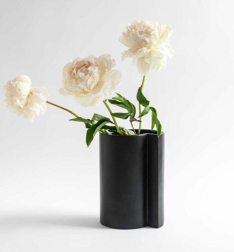 A black Kish vase holds three white peonies with green leaves against a plain Sara Khamisi background.